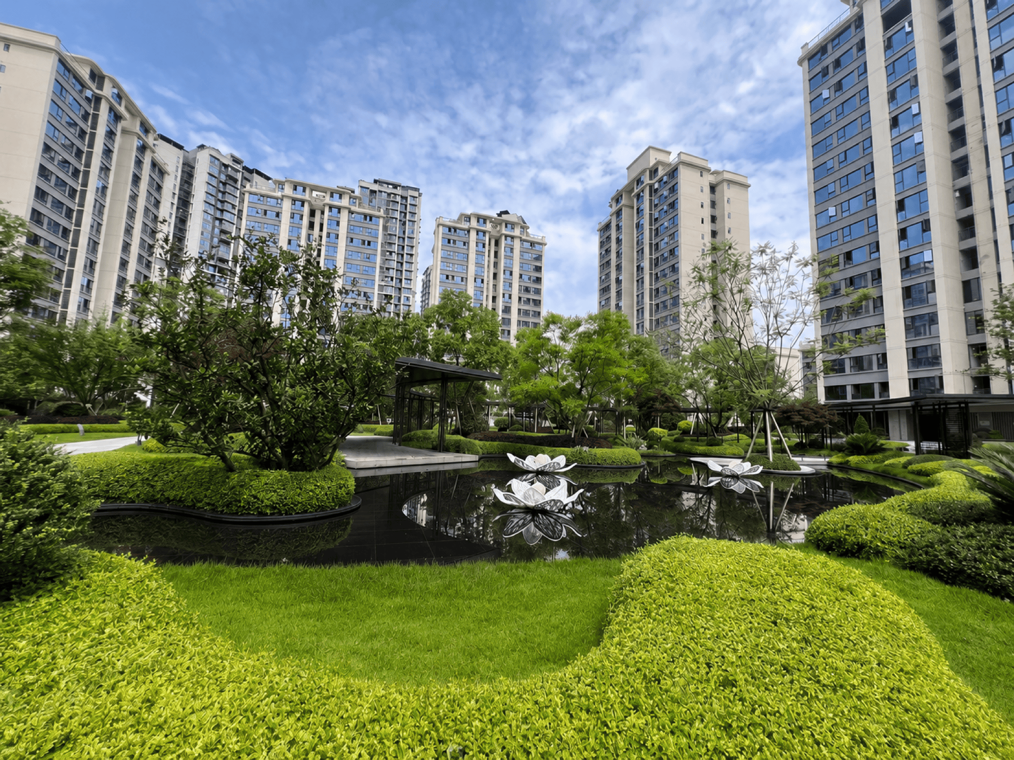 小区中心园林实景：黑色镜面水景与莲花雕塑，环绕修剪整齐的草坪绿植，背景为多栋高层住宅
