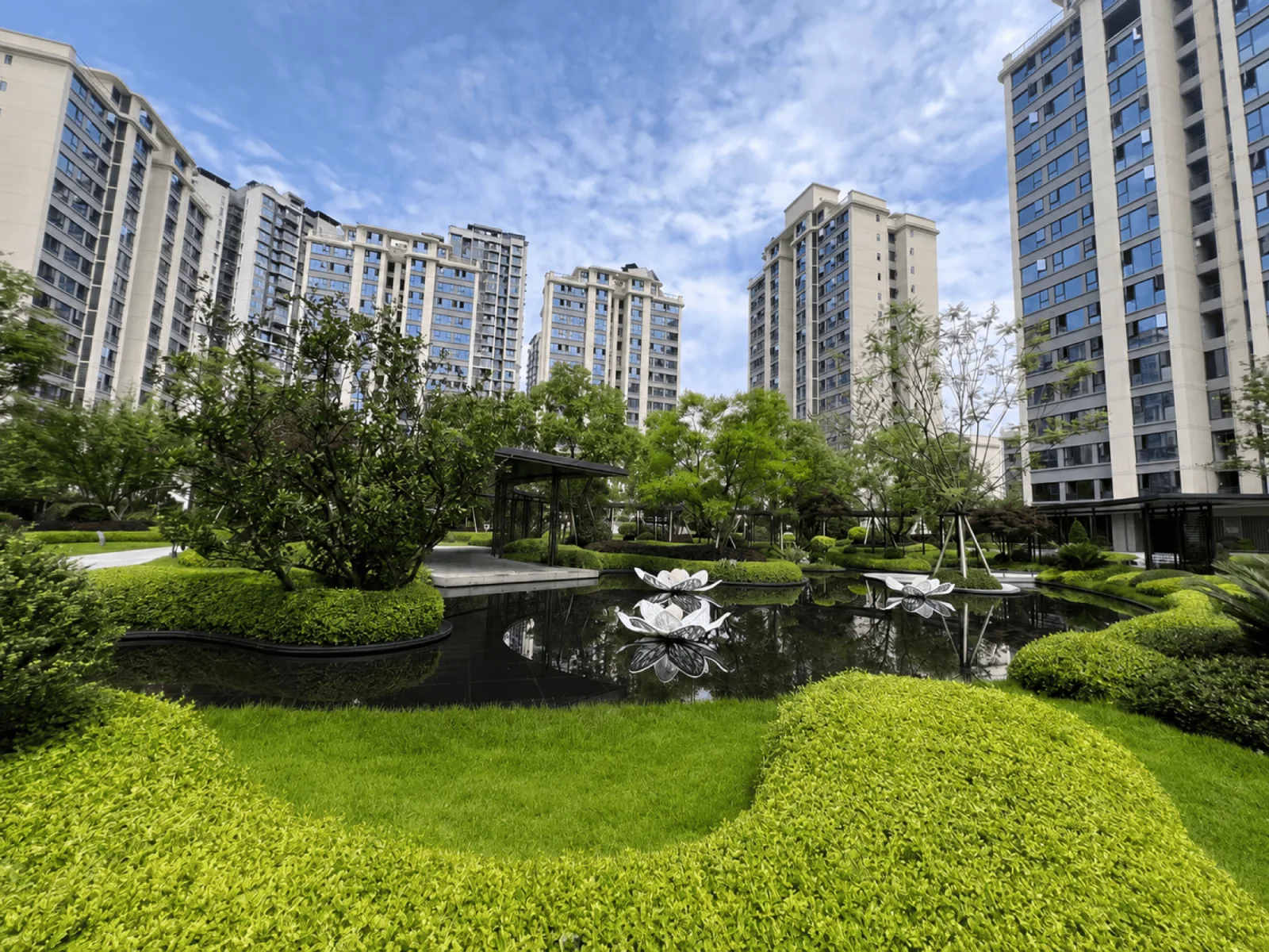 小区中心园林实景：黑色镜面水景与莲花雕塑，环绕修剪整齐的草坪绿植，背景为多栋高层住宅